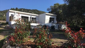 Une maison moderne à deux étages, avec une façade blanche, un balcon et un jardin agrémenté de plantes aux feuilles rouges et bordé de pierres.