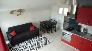 A compact living space with a black sofa, a dining table with chairs, a kitchenette with red cabinets, and a patterned rug. - Le232 fr Meublé 4 Personnes Draps, Serviettes et Ménage Inclus (Chasseneuil-du-Poitou)