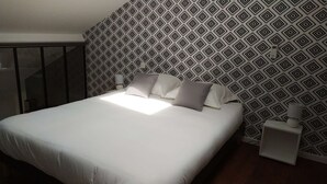 A neatly made bed with white linens and pillows, a bedside table with a lamp, and a patterned wallpaper. - Le232 fr Meublé 4 Personnes Draps, Serviettes et Ménage Inclus (Chasseneuil-du-Poitou)