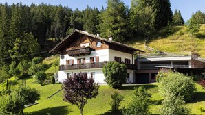 Eine zweistöckige Chalet mit Balkon, umgeben von üppiger Vegetation und einem bewaldeten Hang.