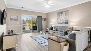 A spacious living room with a grey sofa, a coffee table, and a TV on a stand. There is a sliding glass door leading to a balcony. - Three Bedroom Resort Condo close to Golf, Beach, Shopping & Popular Attractions (Myrtle Beach)