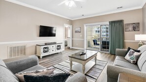 A living room with a flat-screen TV, a ceiling fan, a white cabinet, and a large window with a view of the outside. - Three Bedroom Resort Condo close to Golf, Beach, Shopping & Popular Attractions (Myrtle Beach)