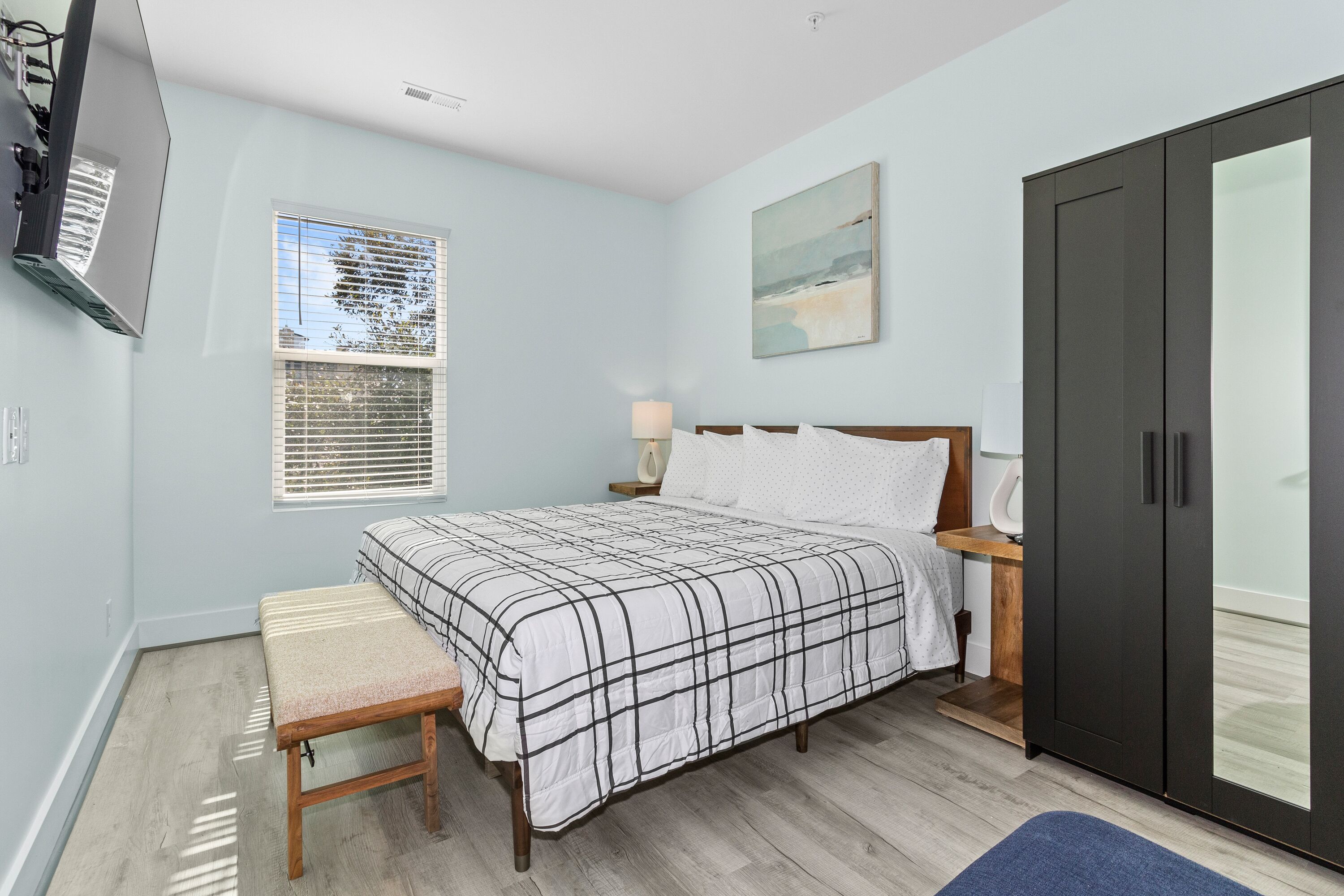 A bedroom with a bed, bench, wardrobe, bedside table, lamp, and a window with blinds.