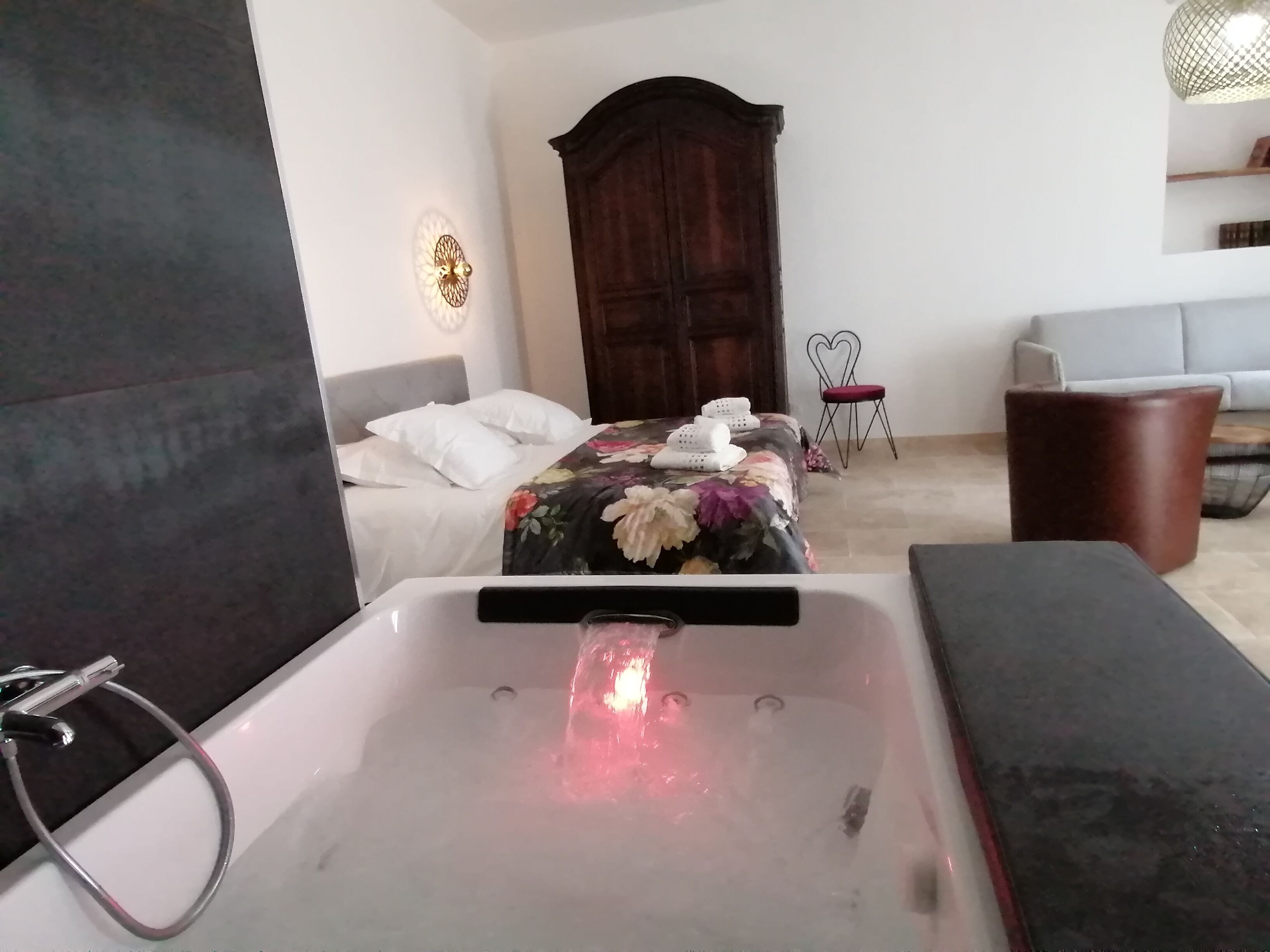 Un dormitorio moderno con una bañera grande, una cama con ropa de cama floral, un armario de madera y una pequeña zona de estar con una silla y sofá.