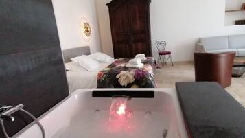 Un dormitorio moderno con una bañera grande, una cama con ropa de cama floral, un armario de madera y una pequeña zona de estar con una silla y sofá.