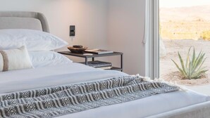 A bedroom with a bed, bedside table, lamp, and a view of the desert through a large window.