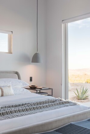 A modern bedroom with a large bed, a bedside table, a lamp, and a view of a desert landscape through a large window.