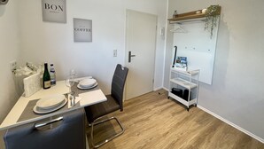 Uma área de refeições com uma mesa posta para dois, uma cadeira, uma prateleira pequena com uma planta e uma porta.