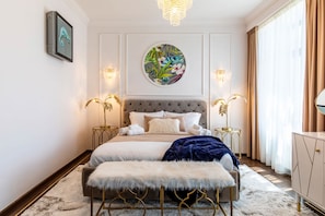 A bedroom with a tufted headboard, a round wall art piece, two bedside tables with lamps, a bench, and a large mirror.