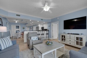 Un salon spacieux avec un canapé gris, une table basse et un téléviseur. Il y a également un coin cuisine avec une table et des chaises, ainsi qu'un ventilateur de plafond.