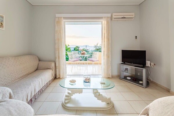 A living room with a sofa, a glass coffee table, a TV on a stand, and a view of the outdoors through a door.