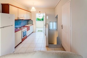 A compact kitchen with a refrigerator, stove, and sink, leading to a balcony with a view of greenery.
