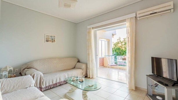 A living room with a sofa, a glass coffee table, a TV on a stand, and a balcony with a view.