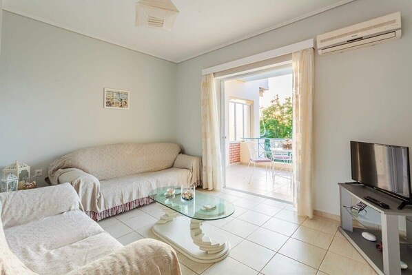 A living room with a sofa, a glass coffee table, a TV on a stand, and a balcony with a view.