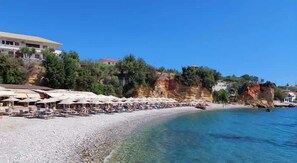 A beach with umbrellas and lounge chairs, a rocky cliff, and buildings in the background.