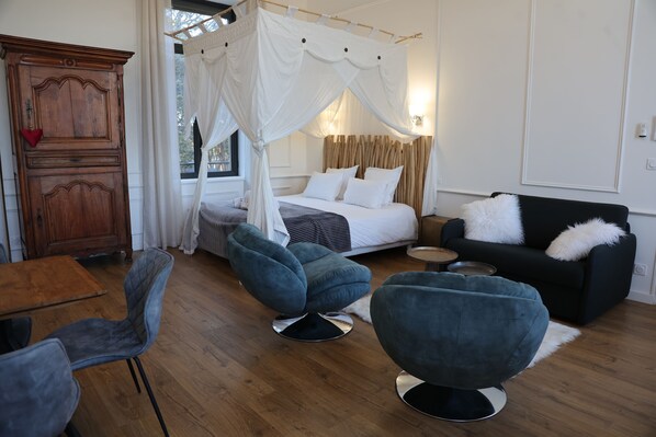A bedroom with a canopy bed, a wooden wardrobe, a dining table with chairs, and a black sofa with white cushions.