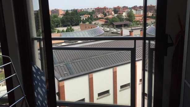 A view through a window of a building with multiple floors, featuring a mix of residential and commercial buildings in the background. - appartamento luminoso  con balcone, silenzioso davanti a un parco vicino a GYM (Gallarate)