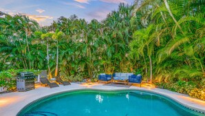 A swimming pool surrounded by lush greenery and palm trees, with lounge chairs and a barbecue grill. - Chic Pool Home Steps to Beach! Game Room and Paddleboards (Holmes Beach)