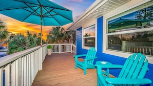 A wooden deck with turquoise Adirondack chairs and a matching umbrella, overlooking a blue house with a large window and a view of palm trees. - Chic Pool Home Steps to Beach! Game Room and Paddleboards (Holmes Beach)