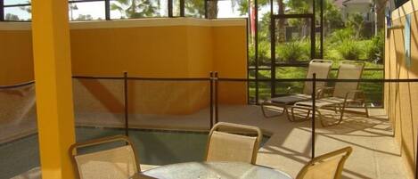 Un espacio de esparcimiento al aire libre con mesa y sillas de vidrio, rodeado por un muro amarillo y una barrera de cristal.