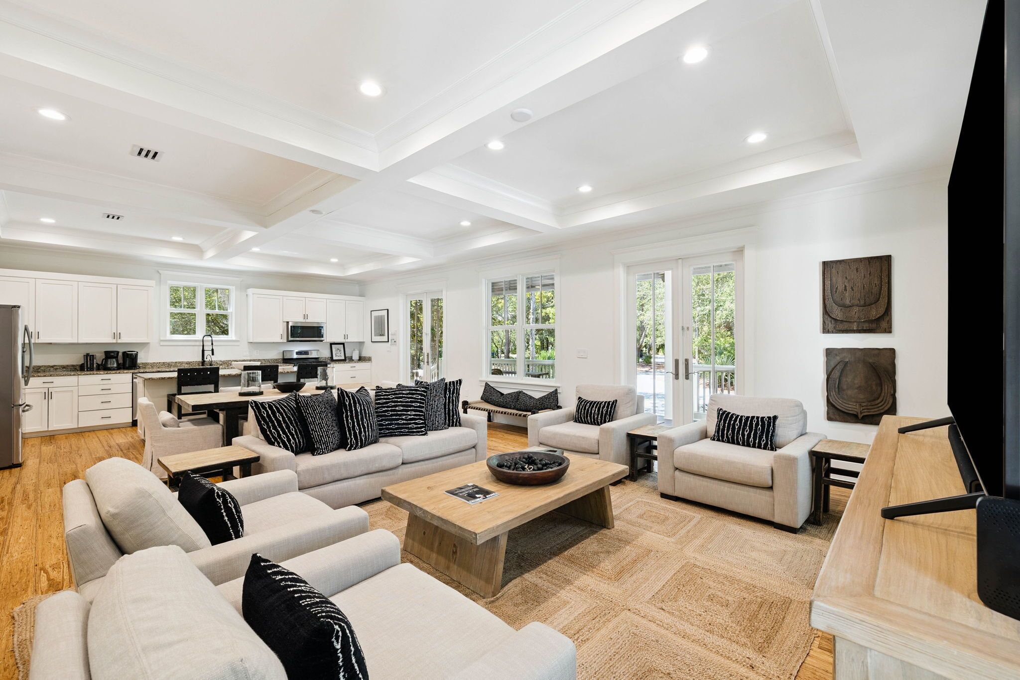 A spacious living area with a large sofa set, a coffee table, and a television. There is a kitchen area with white cabinets and a dining table.