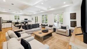 A spacious living area with a large sofa set, a coffee table, and a television. There is a kitchen area with white cabinets and a dining table. - Charming home perfectly located near Seaside, Watercolor, and the best of 30A (Seagrove Beach)