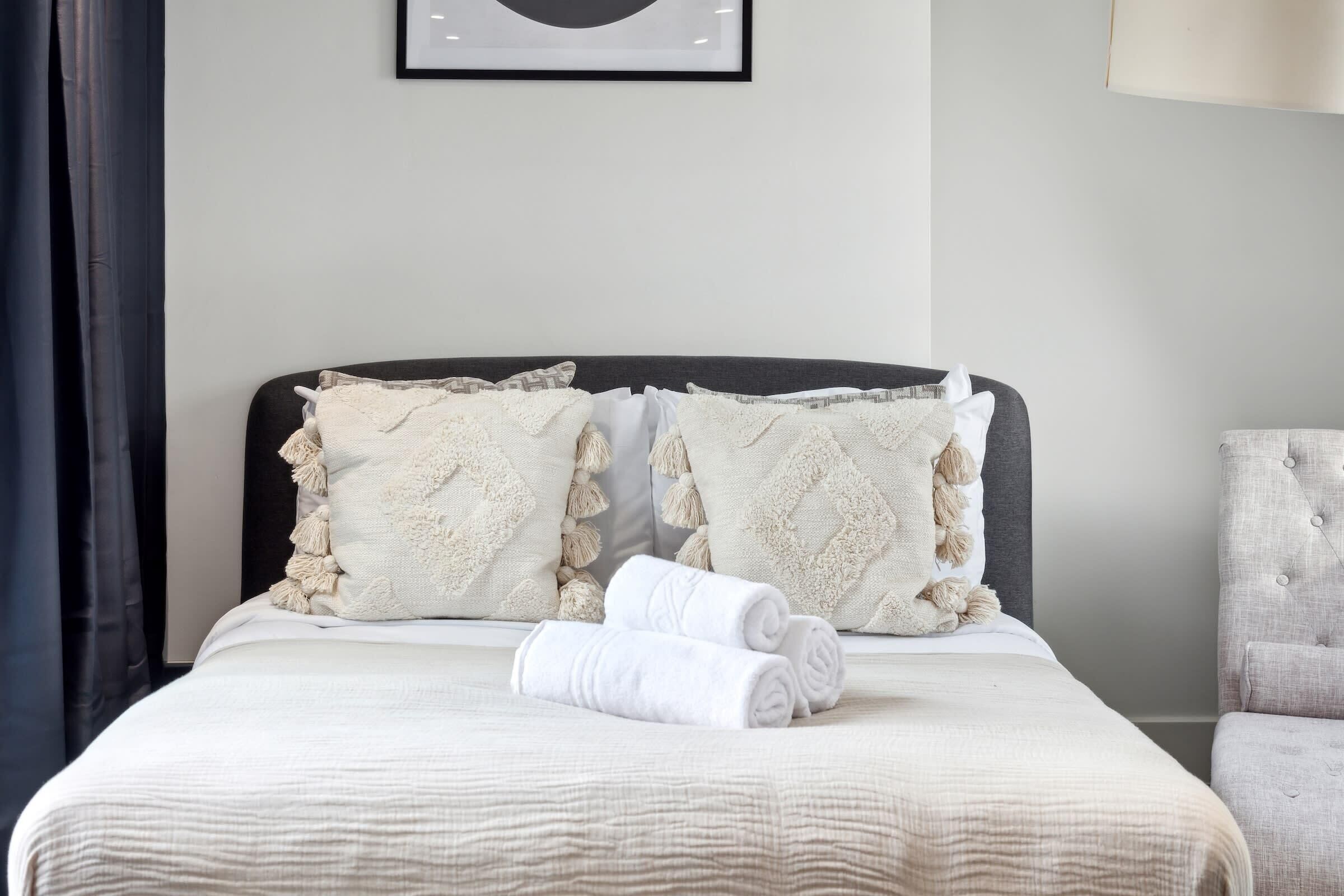 Una cama bien hecha con edredón blanco, dos toallas blancas enrolladas y almohadones decorativos con borlas.