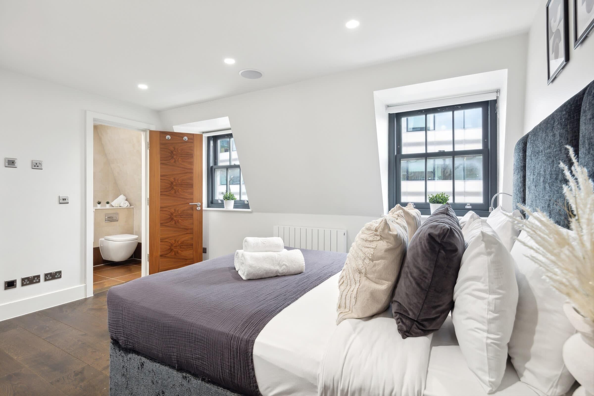 A modern bedroom with a large bed, a wooden door leading to a bathroom, and a window with blinds.