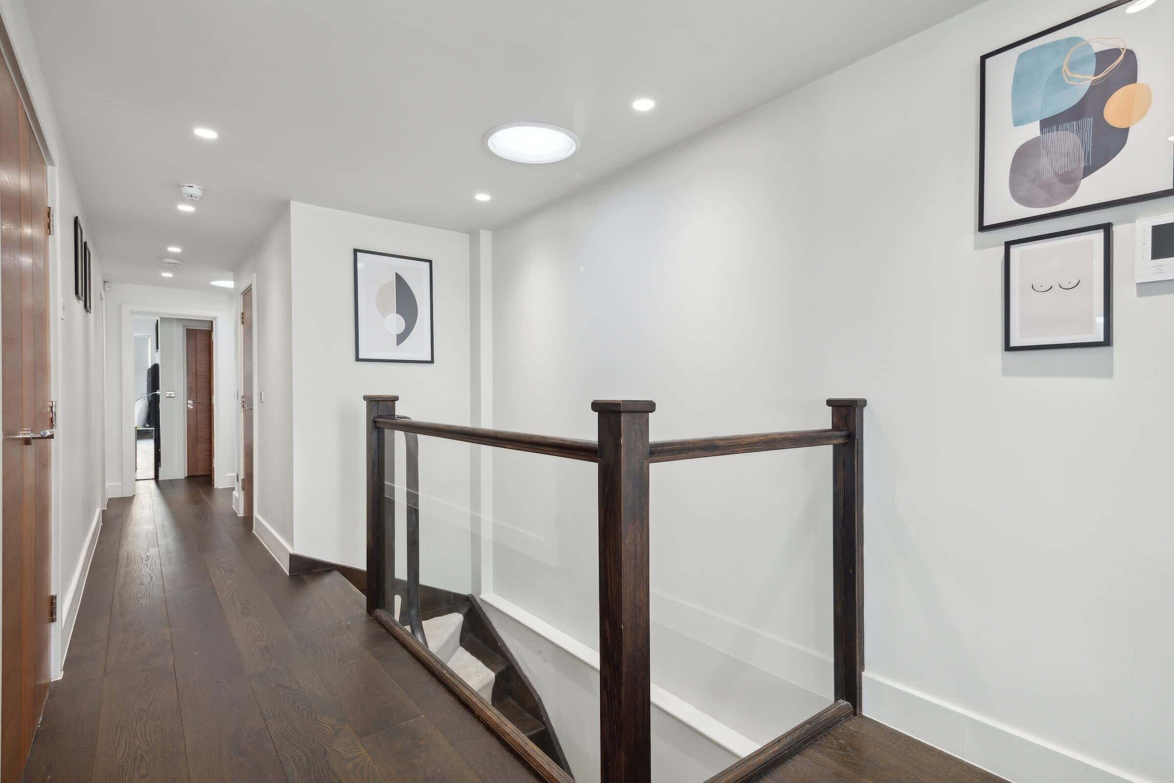 A modern hallway with wooden flooring, a glass railing, and artwork on the walls.