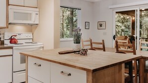 Una cocina moderna con armarios blancos, microondas, estufa y una mesa de comedor de madera con sillas. Hay ventanas con vistas a árboles y una lámpara en el techo.