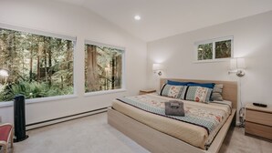 Un dormitorio con una cama grande, dos mesitas de noche, una silla y vistas a un bosque a través de amplias ventanas.