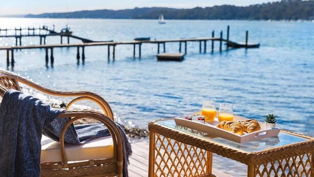 A wicker chair with a cushion and a tray table with croissants, juice, and a small plant. - Scotland Island Waterfront House w/ Private Jetty (Scotland Island)