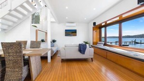 A modern interior with a wooden floor, a sofa, a dining table with wicker chairs, and a large window overlooking a body of water. - Scotland Island Waterfront House w/ Private Jetty (Scotland Island)