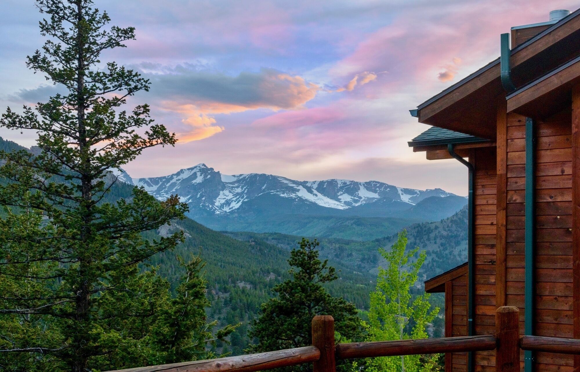 A wooden cabin with a view of snow-capped mountains and a forested valley.