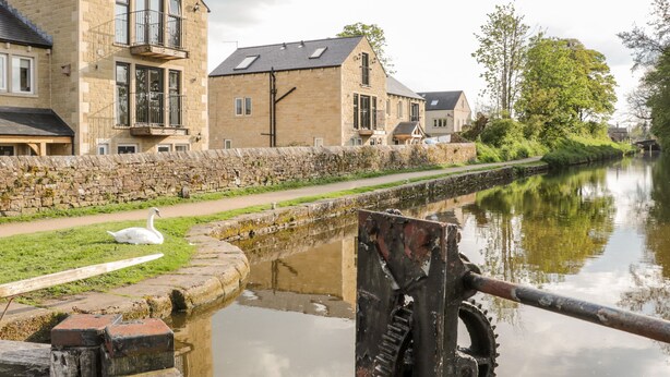 A canal with a swan, stone buildings, and a water wheel. - 6 Lock View (Skipton)
