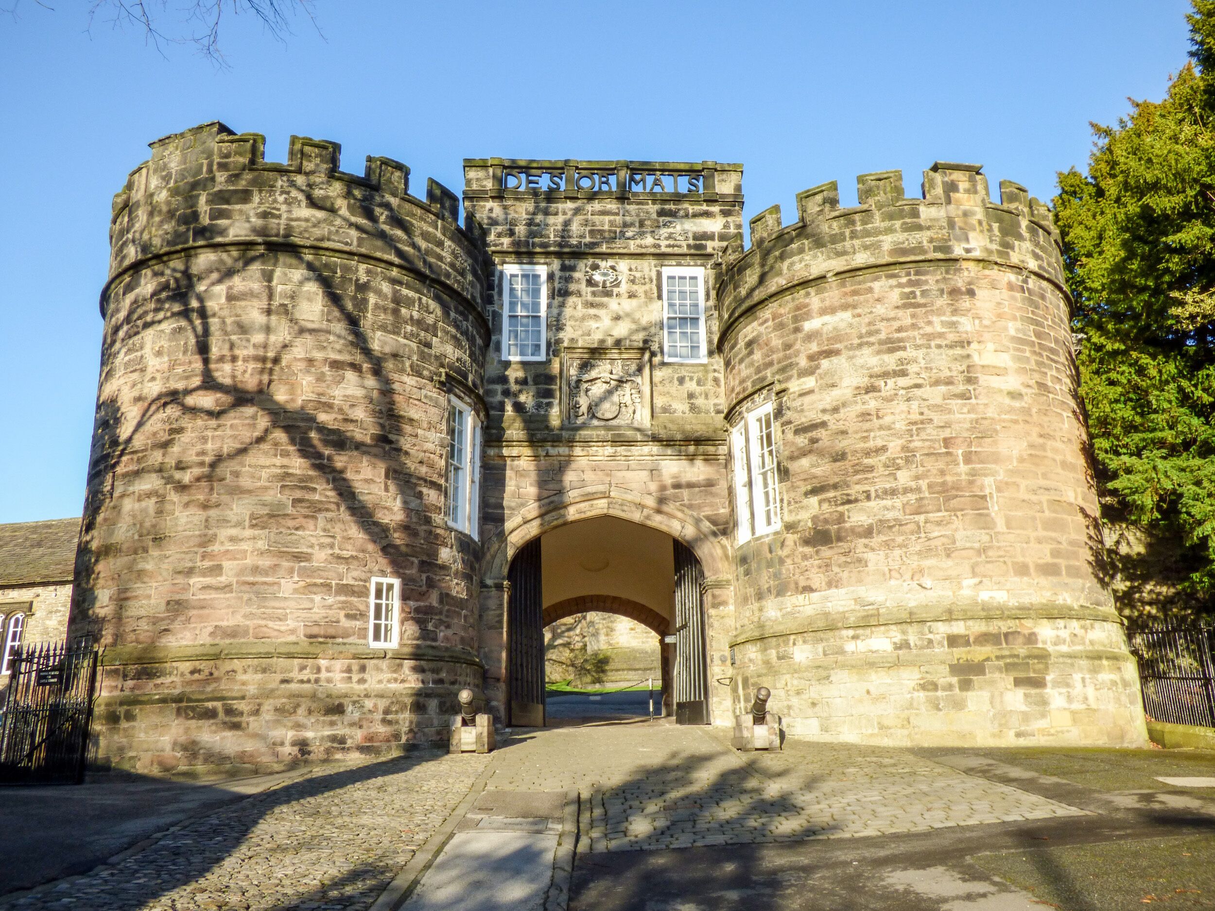 A historic stone building with a central archway and two round towers, featuring a sign that reads "DES OF TAI."