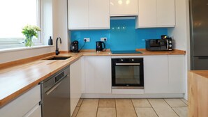 A modern kitchen with white cabinets, a blue backsplash, a dishwasher, an oven, and a refrigerator. - Estuary View (Newquay)