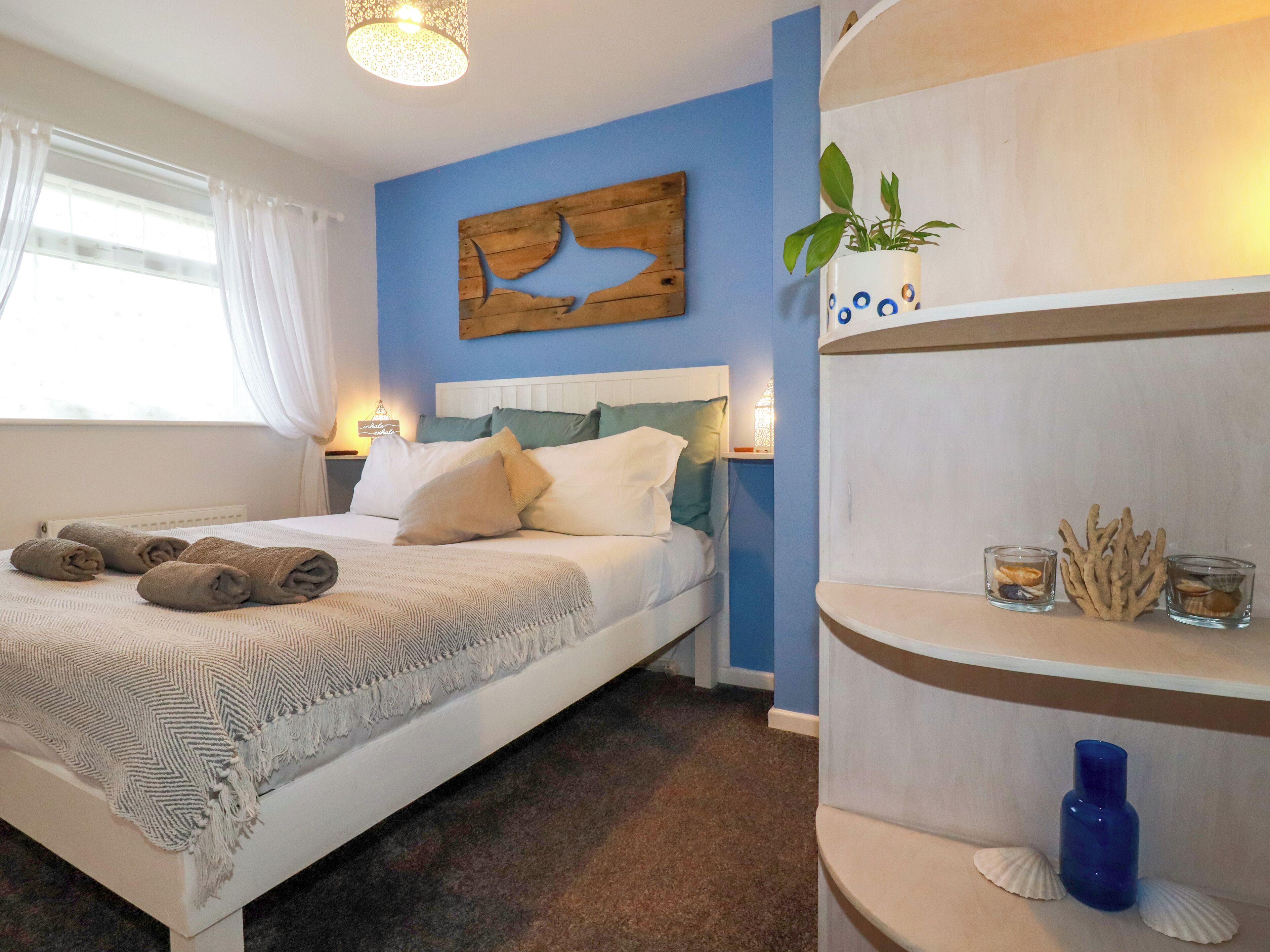 A neatly made bed with rolled towels, a blue accent wall, and a shelf with decorative items.