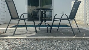 An outdoor patio with a table and four chairs, a pool, and a white building with a sliding glass door.