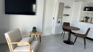 A modern living room with a flat-screen TV, a sofa, a dining table with chairs, a small side table with a plant, and a kitchen area with a microwave and utensils.
