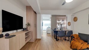 Un salon moderne doté d’un téléviseur à écran plat, de meubles en bois, d’un coin repas avec une table et des chaises, et d’un canapé agrémenté d’un plaid orange.