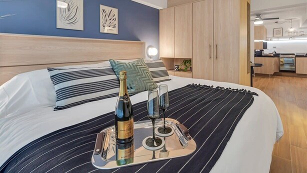 Une chambre d’hôtel moderne, équipée d’un lit, d’une table de chevet avec une bouteille et des verres, et offrant une vue sur la cuisine.