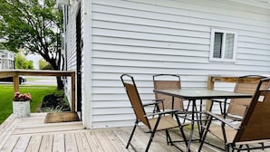 Une terrasse en bois avec une table et des chaises, une plante en pot et une maison aux murs recouverts de bardage blanc.