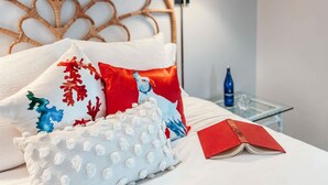 Una cama bien hecha con varios almohadones, un libro y una botella sobre una mesita de noche.