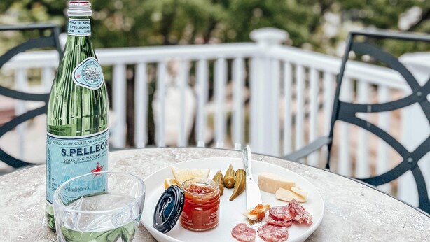 Una mesa con una botella de agua con gas San Pellegrino, una copa, y un plato con queso, aceitunas y salami.