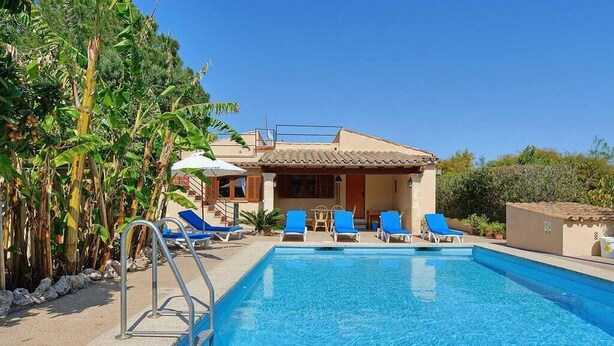 A swimming pool with blue water, surrounded by lounge chairs and a building with a tiled roof.