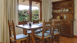 A dining area with a wooden table and chairs, a bookshelf, and a view of greenery through a window.