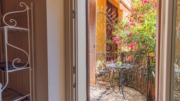 A narrow outdoor corridor with a metal chair and table, a metal shelf unit, and a door leading to a garden with blooming flowers.