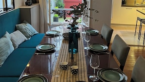 Un coin repas avec une longue table dressée pour six personnes, un canapé et une vue sur un bâtiment rouge à l’extérieur.