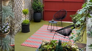 Une petite terrasse extérieure avec un tapis à carreaux, des plantes en pot et, au fond, une porte de garage rouge.
